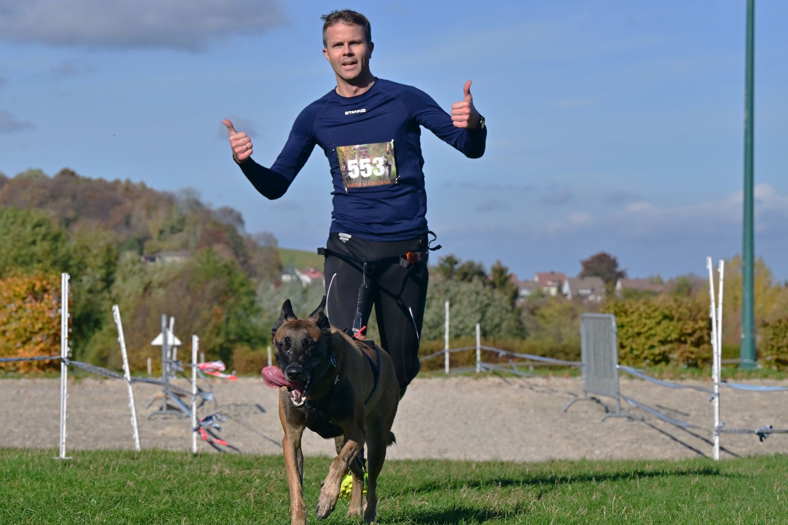 Coureur en haut bleu marine à manches longues, avec un dossard, pouces levés, et son chien au pelage foncé qui court devant lui sur un terrain de sable délimité par des barrières, sous un ciel bleu