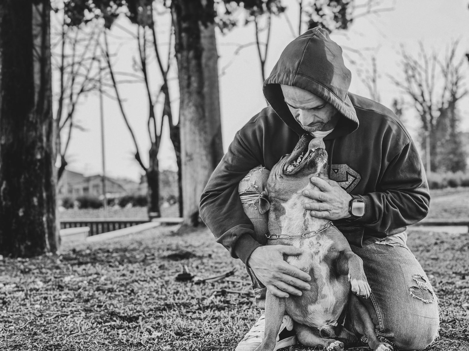 Photo en noir et blanc : un homme en sweat à capuche, à genoux dans un parc, serre son chien contre lui, le museau du chien tout près de son visage