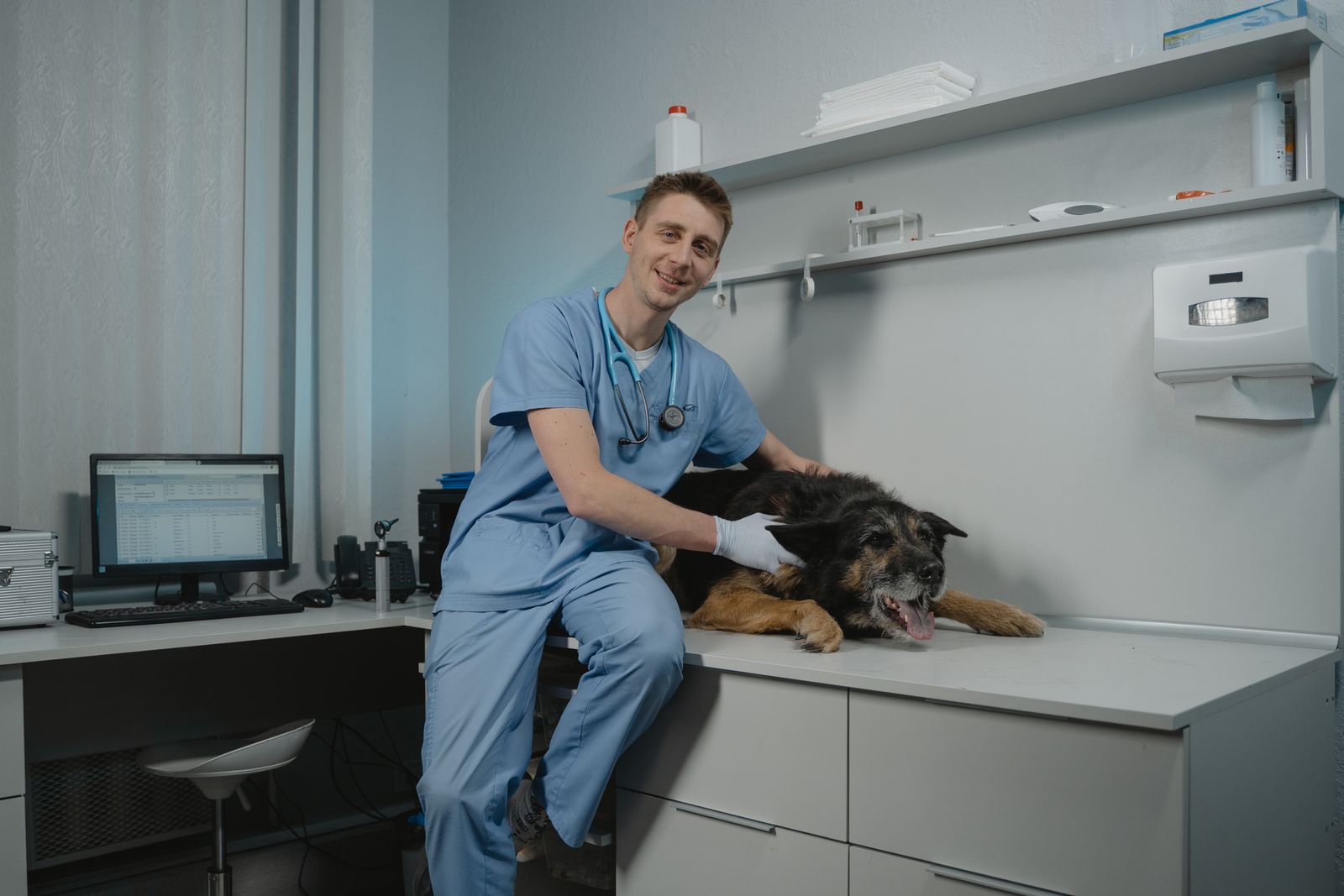 Vétérinaire en tenue bleue, stéthoscope autour du cou, assis sur la table d'examen, la main gantée posée sur un grand chien noir et feu couché