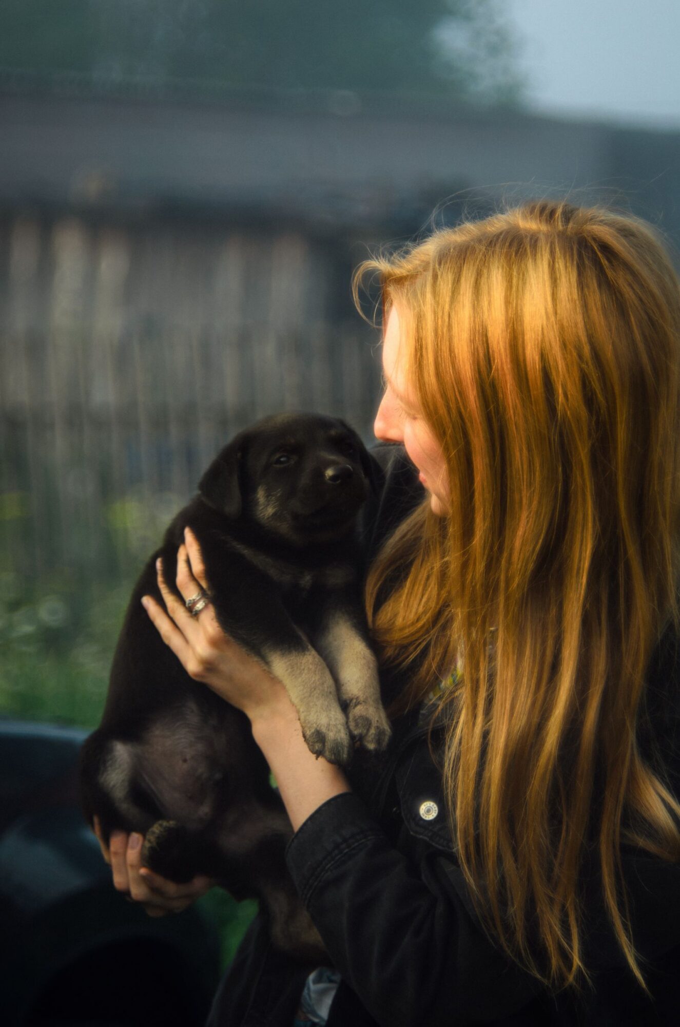 Femme aux longs cheveux blond vénitien, vêtue de noir, qui tient dans ses mains un chiot noir aux pattes claires, devant une clôture en bois floue