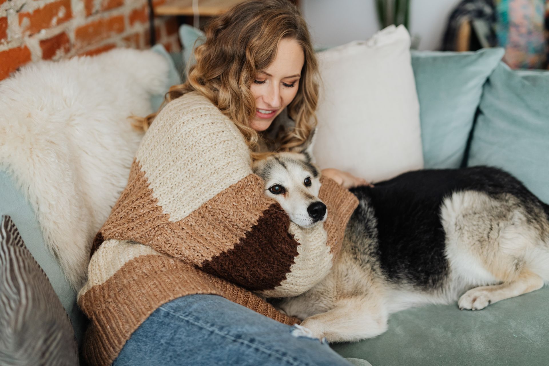 Femme aux cheveux blonds ondulés, vêtue d'un gros pull en tricot beige et marron, qui sourit en regardant son chien sur un canapé vert d'eau ; le chien au poil noir, blanc et fauve, couché contre elle, pose la tête sur le pull et regarde l'objectif